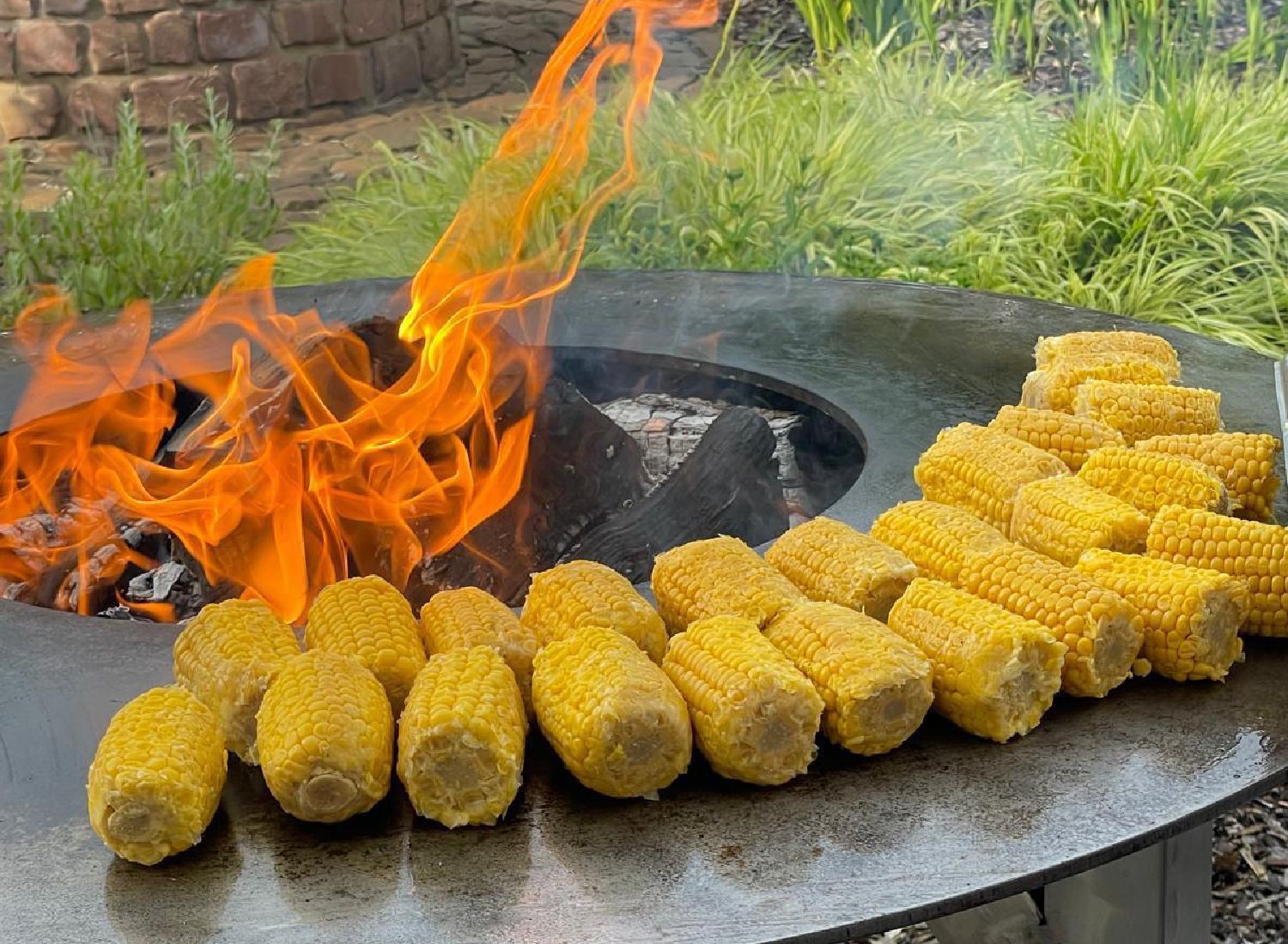 Maiskolben werden am Rand eines brennenden Feuers auf einem Tisch gezeigt.